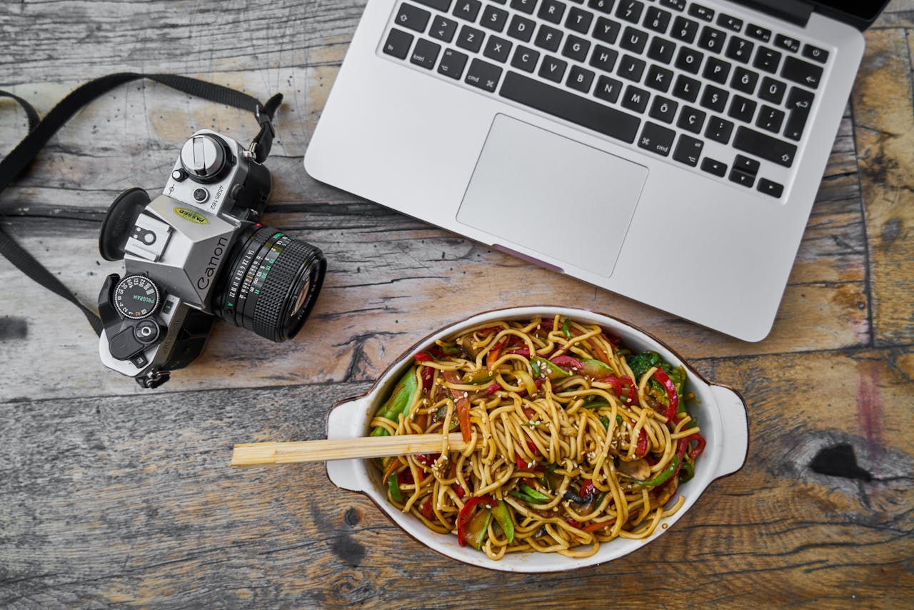 Top view of a film camera, laptop, and a bowl of Asian noodles on a rustic table.