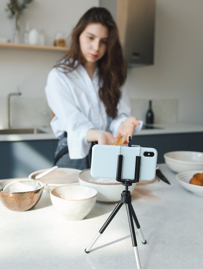 Young woman vlogging her cooking session in a modern kitchen using a smartphone and tripod.