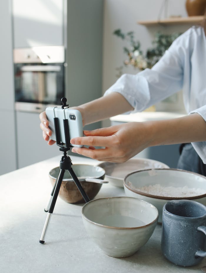 A person records a cooking process in a kitchen using a smartphone and tripod.